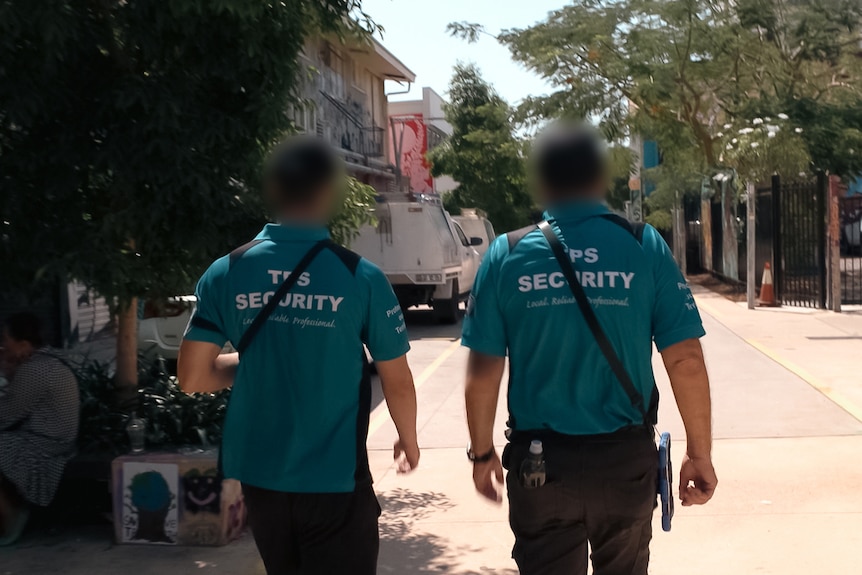Two men wearing caps and  blue shirts with 'TPS security' walk away from the camera down a city laneway. People sit nearby.
