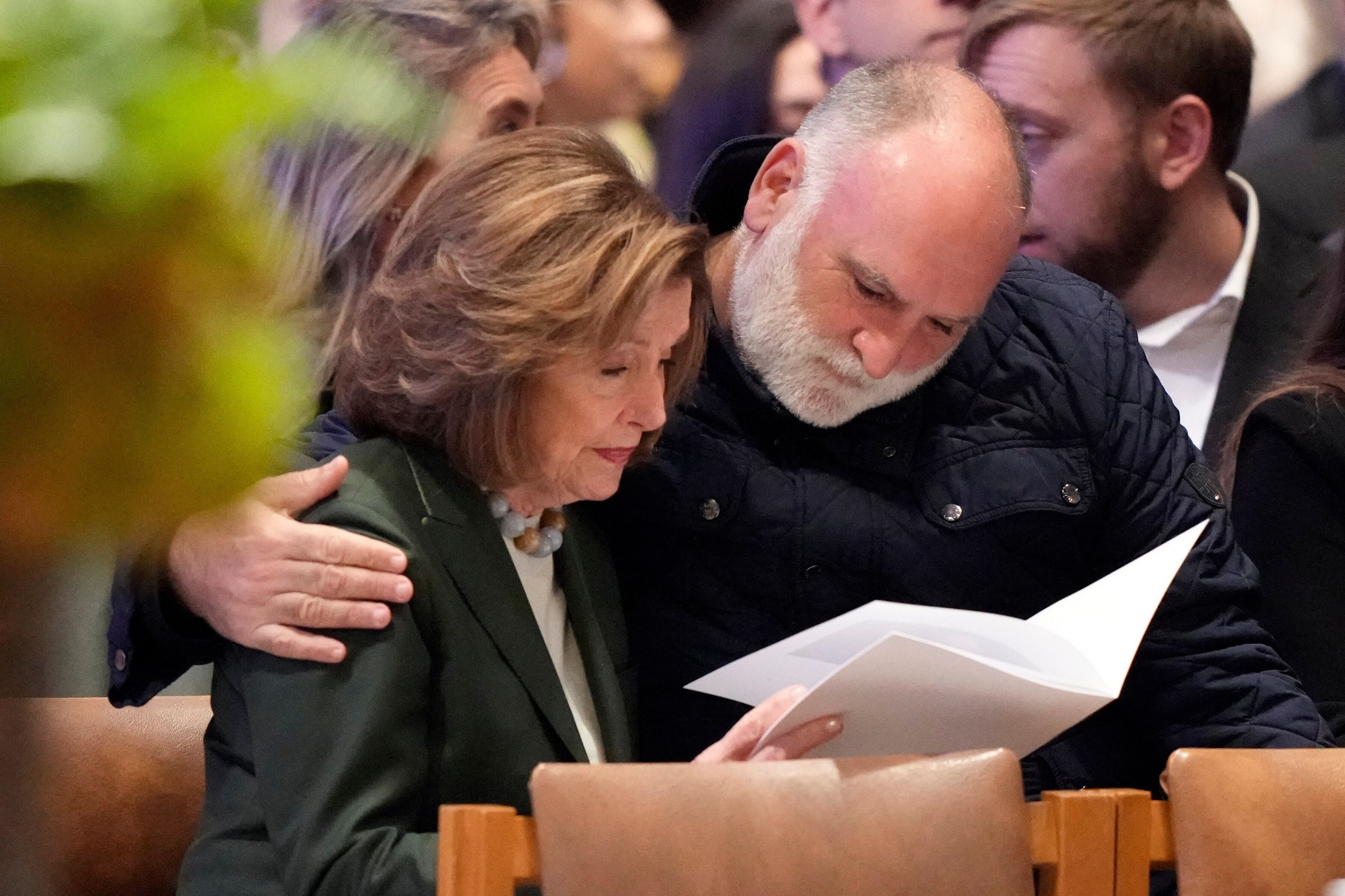 An older woman reads a booklet while a bearded man puts his arm around her. They are seated on a pew in a cathedral.