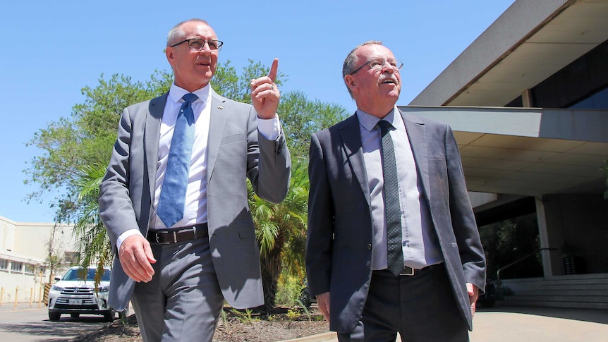 An upshot of two middle aged men in suits looking into the middle distance. One is pointing