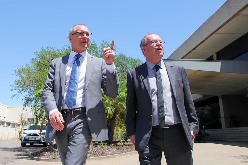 An upshot of two middle aged men in suits looking into the middle distance. One is pointing