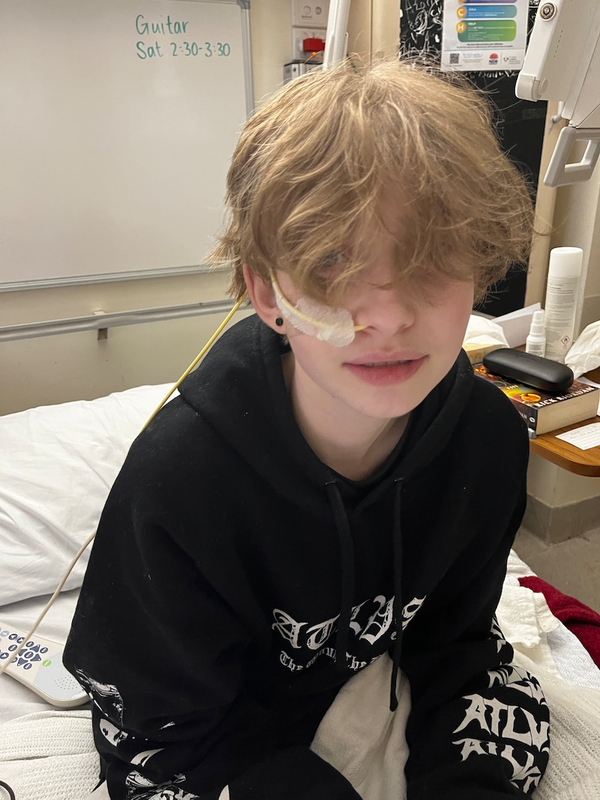 A boy with hair covering his eyes sits on a hospital bed. A feeding tube is coming out of his nose, and is taped to his cheek.