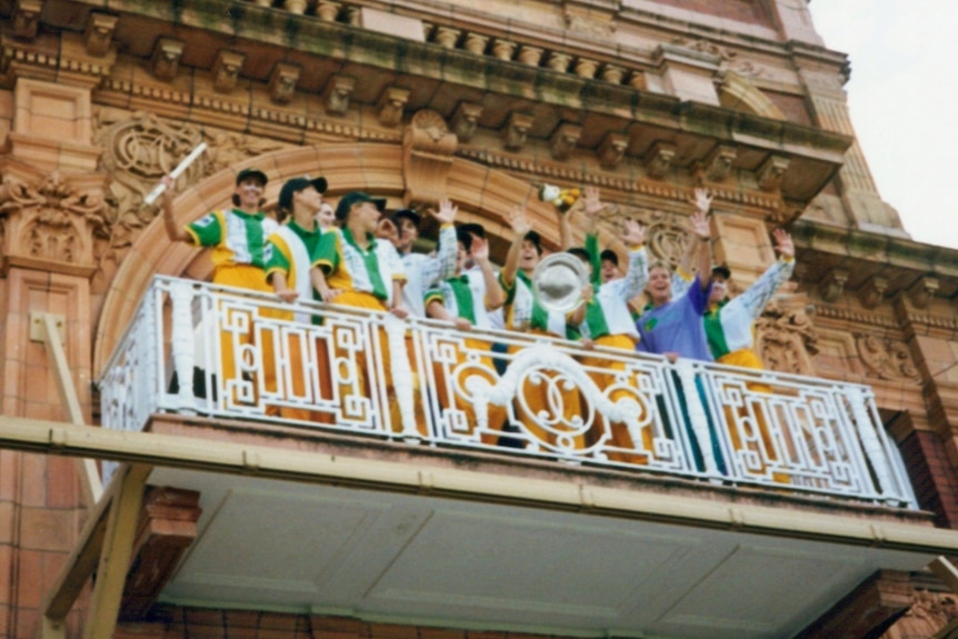 Members of the Australian women's cricket team stand on a balcony holding a trophy, waving and smiling.
