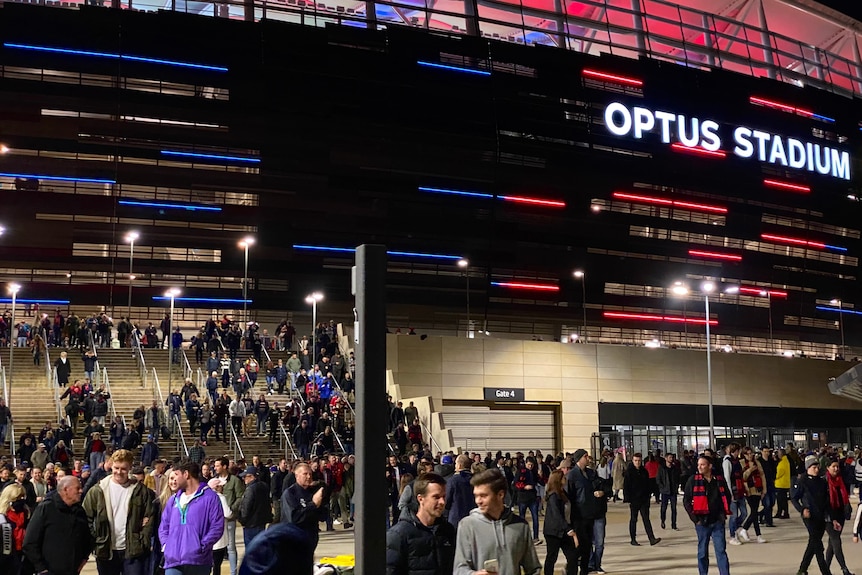 Hundreds of football fans walk down stairs and out of Perth Stadium lit up blue and red at night.