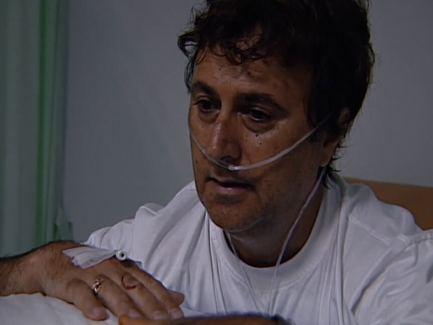 Joe Giardina in hospital with an oxygen tube on. He wears a white t-shirt and looks down solemnly