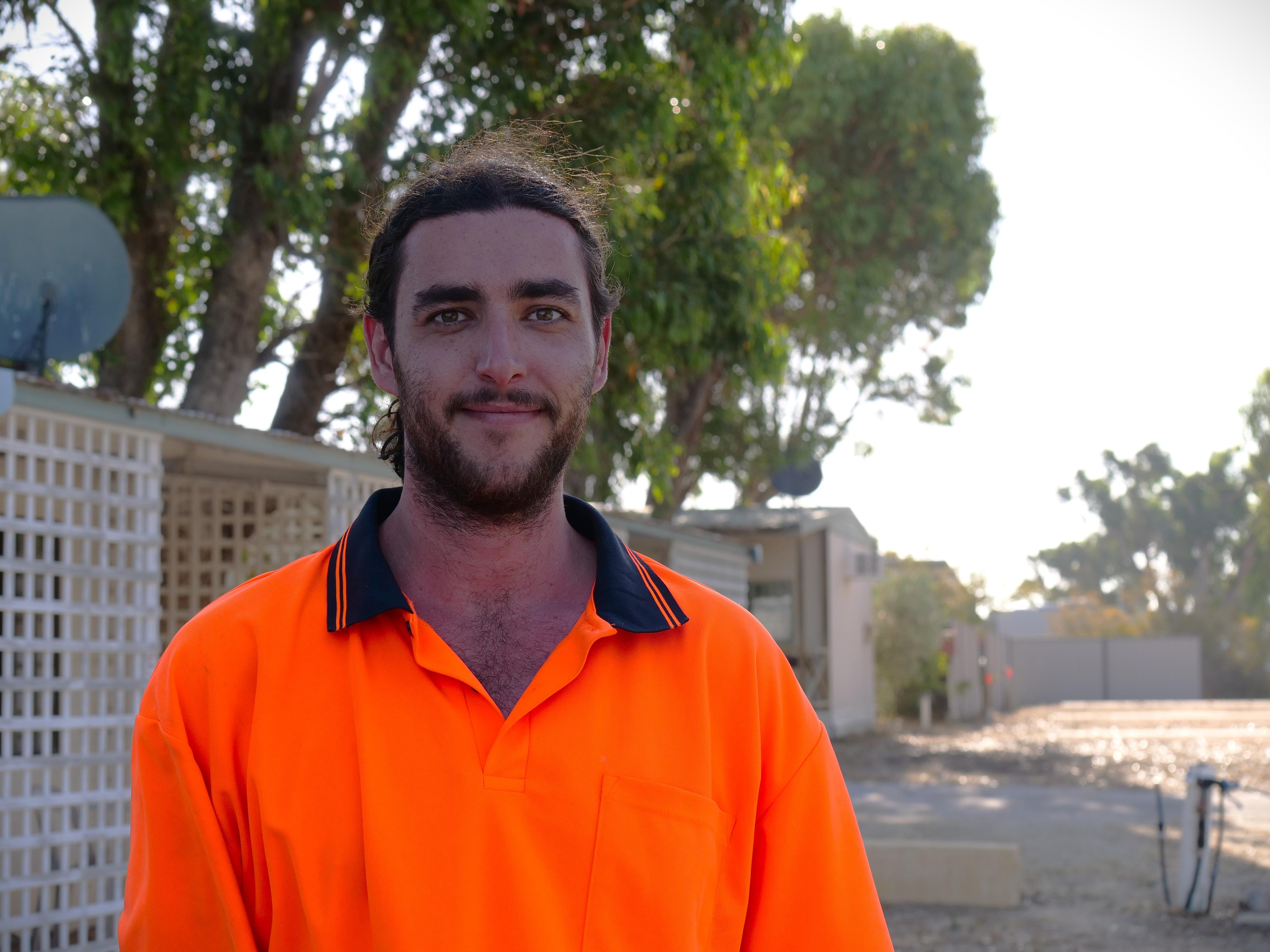 Un joven con barba y cabello recogido, vestido con una camisa de alta visibilidad, mira a la cámara frente a una cabaña. 