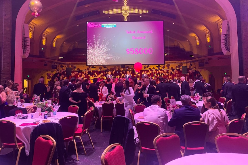 Pink lighting illuminates a charity fundraising event, with tables and chairs in the foreground. 