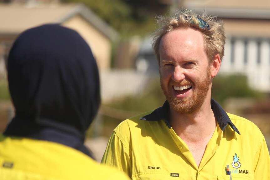 A man in a high-vis shirt with blond hair and a short beard laughs. The back of a woman's head is in the foreground. woman