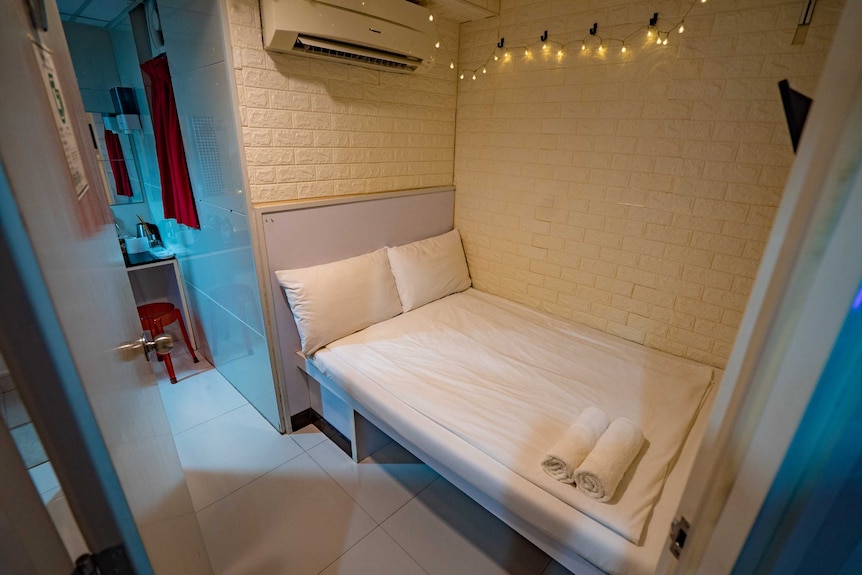 A crammed hotel room with a double bed and bathroom facilities in the corner. There are towels on the bed and fairy lights above