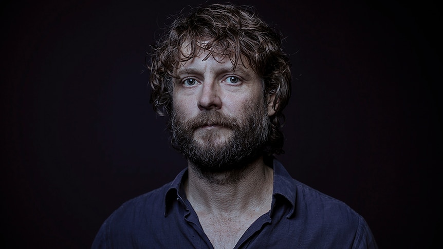 A man with dark brown curly hair and beard stands against black background with serious expression.