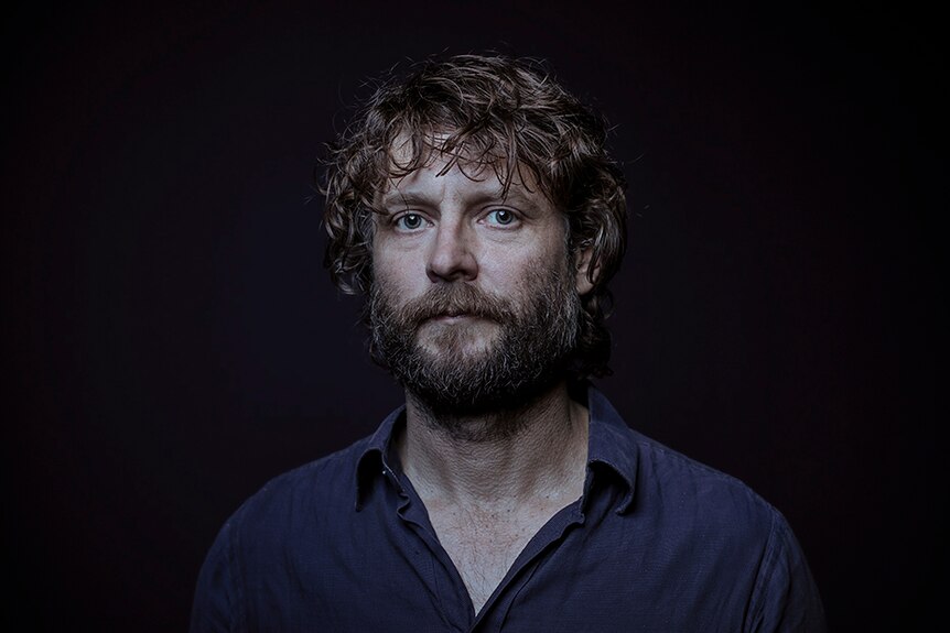 A man with dark brown curly hair and beard stands against black background with serious expression.