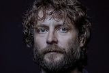 A man with dark brown curly hair and beard stands against black background with serious expression.