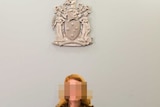 A woman sits under a coat of arms with a blurred face