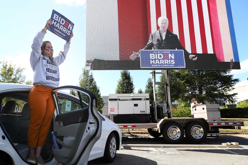 A woman holds up a 'Biden Harris' sign while standing on the footwell of a car at a drive-in campaign rally