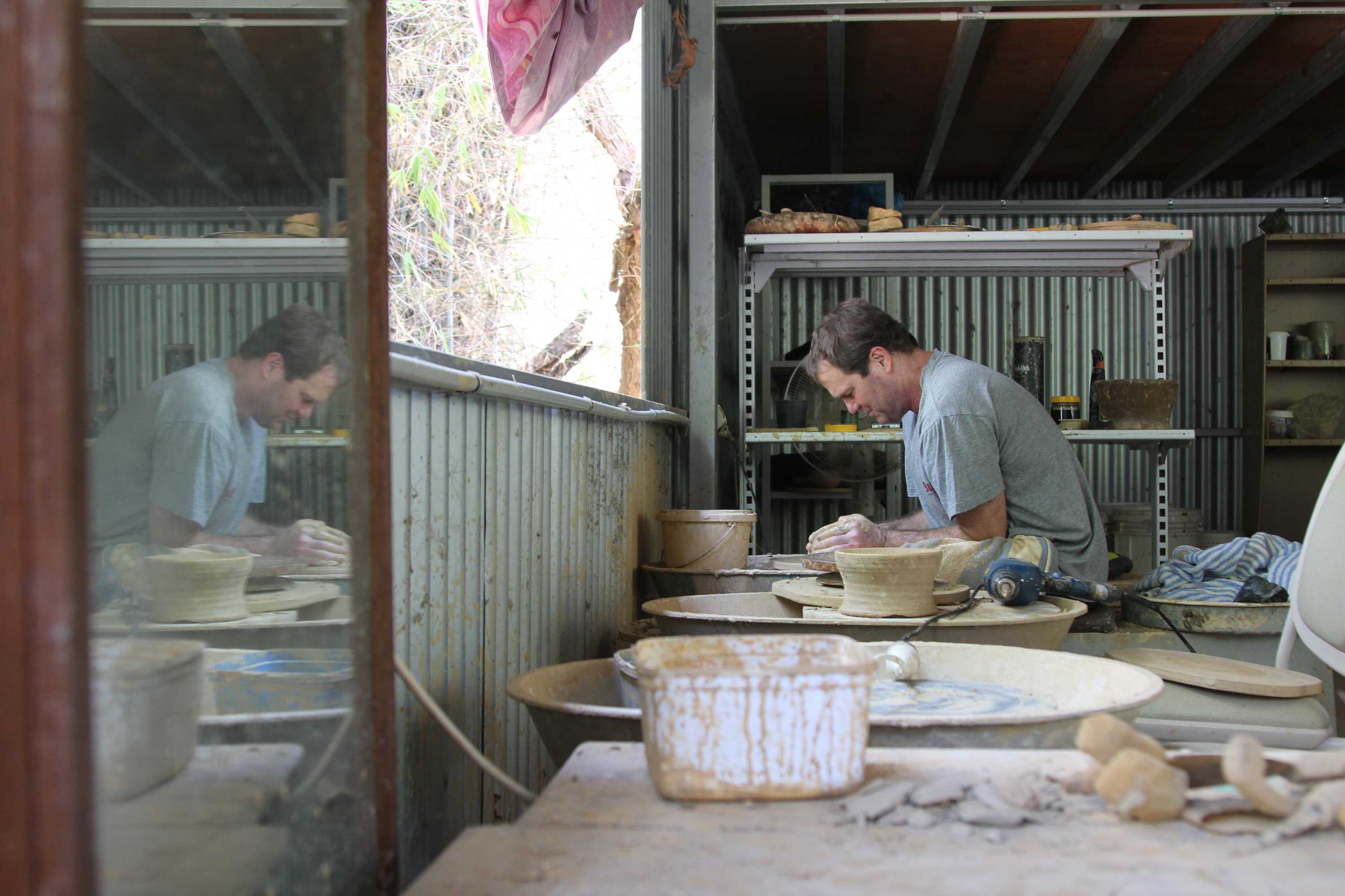 A man sits at a pottery wheel in a shed.