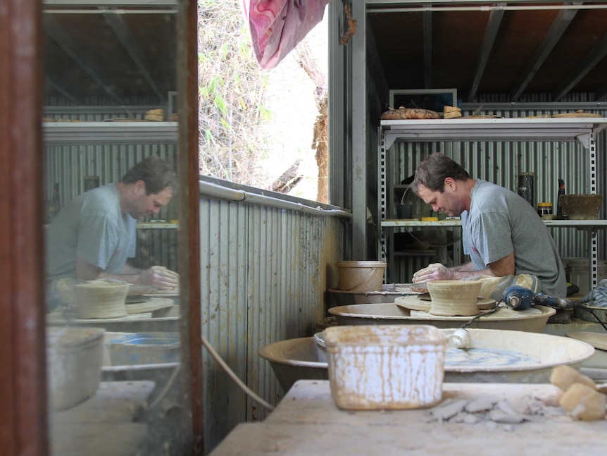 A man sits at a pottery wheel in a shed.