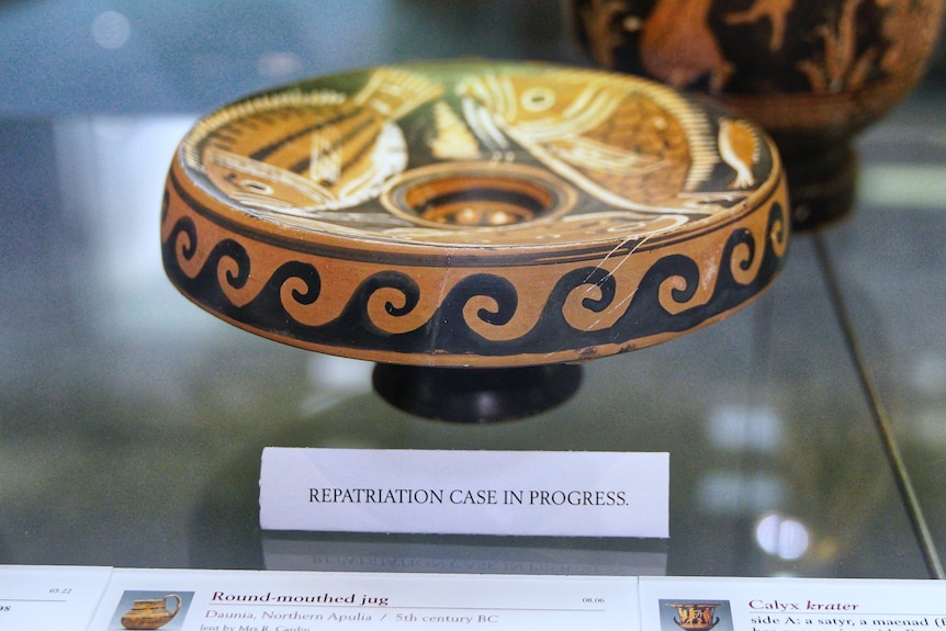 An ancient plate with wave design edges behind a glass display with a sign underneath that reads "reparation case in progress".