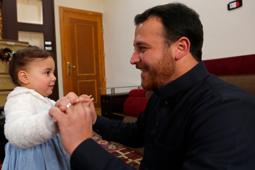 A man in a black jumper plays with a small girl wearing a blue dress and white cardigan.