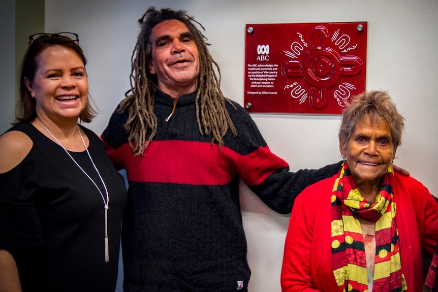 Two women and a man who are Indigenous Australian stand in front of a plaque acknowledging Indigenous ownership of the country.