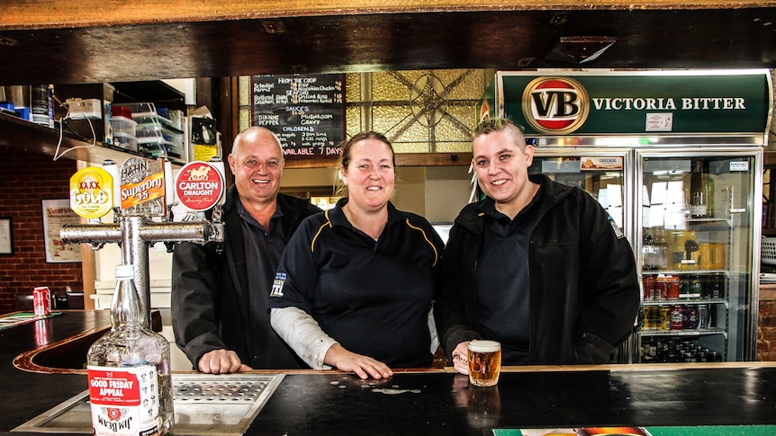 Three peopel standing behind a country pub's bar, with a glass of beer on the bar.