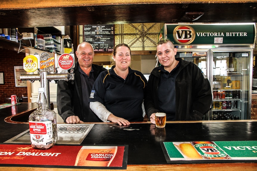 Three peopel standing behind a country pub's bar, with a glass of beer on the bar.