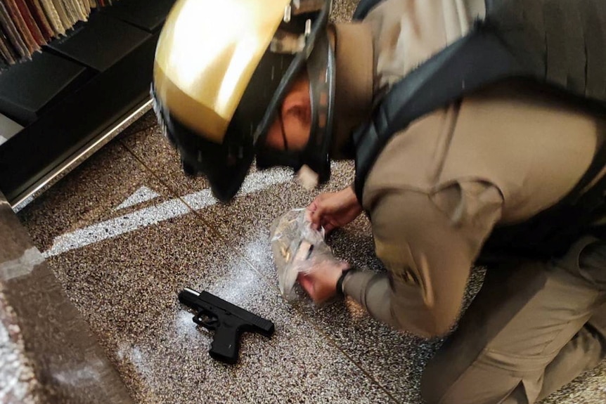A close of an armed man kneeling on the floor near a small gun, which sits on the ground