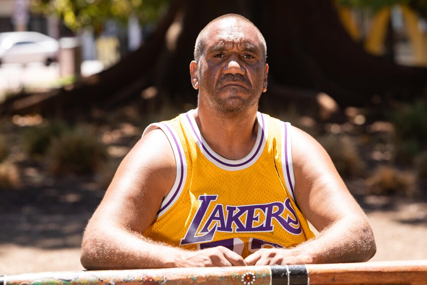 A man in a yellow basketball jumper sits on a park bench, looking at the camera with an unhappy expression.