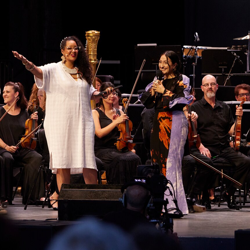 Stéphanie Kabanyana Kanyandekwe in front of an orchestra with an arm outstretched.2 singers hold microphones, the conductor clap