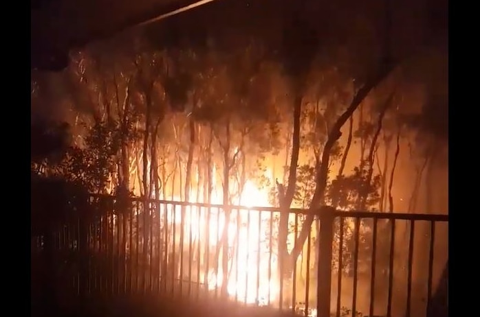 The orange glow of a bushfire can be seen behind the fence line of a home, burning through bushland.