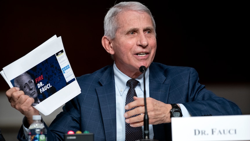 A man with silver hair and a blue suit speaks into a microphone.