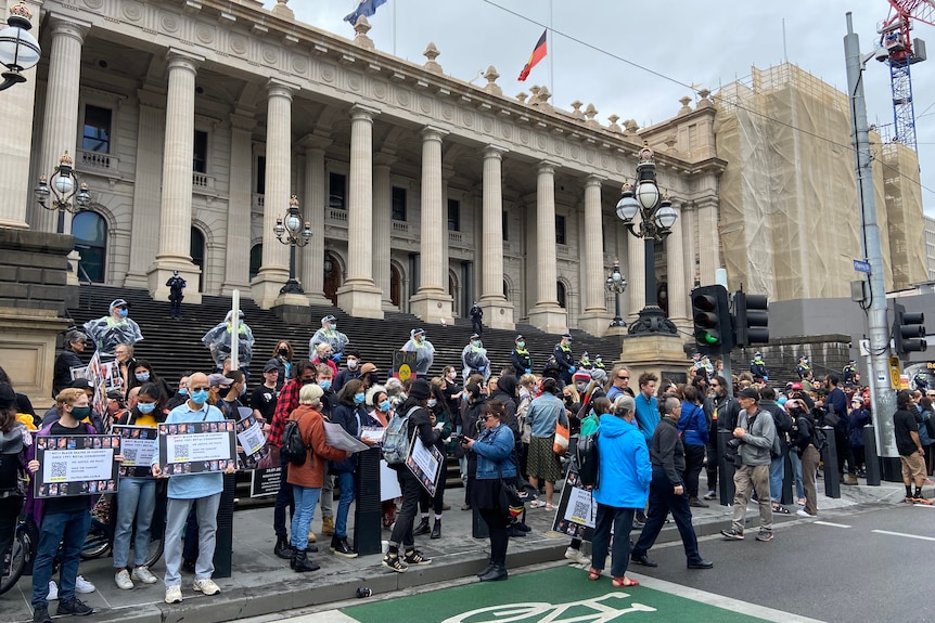 A large group holding protest signs in front of the steps of Parliament House in Melbourne's CBD.