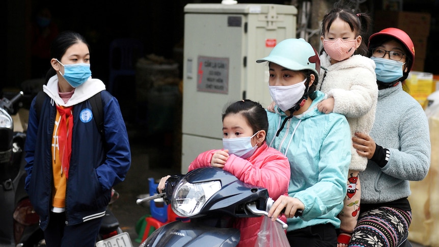 두 명의 여성과 두 명의 어린이가 같은 오토바이를 타는 동안 다른 여성이 거리에서 그들을 지켜보고 있습니다.