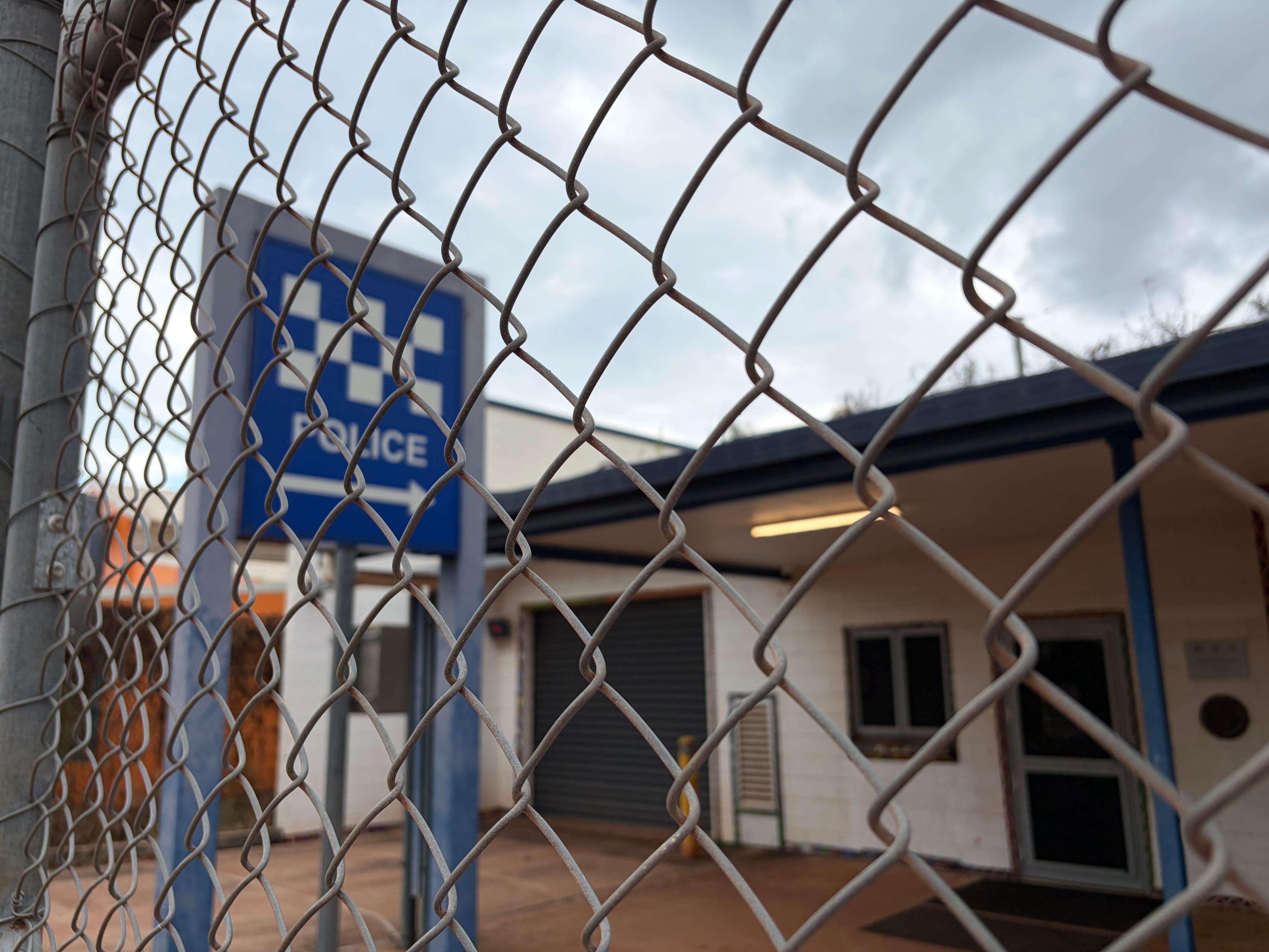 A blue and white chequered sign outside the Wadeye police station as seen through the facility's chain-link fence.