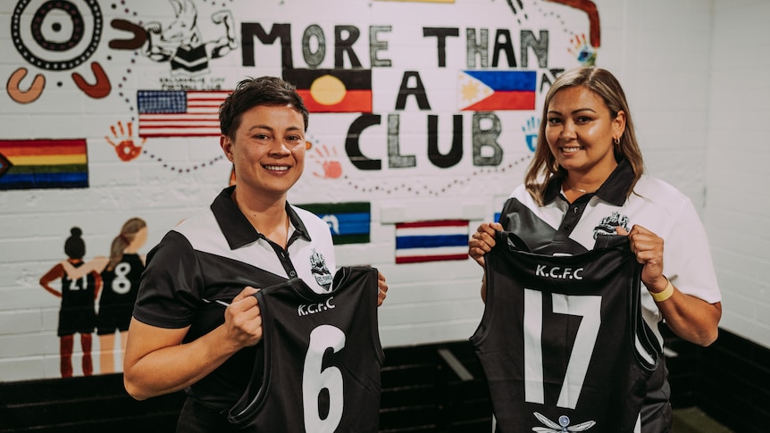 Two women holding Australian Rules football guernseys for the club they represent.  