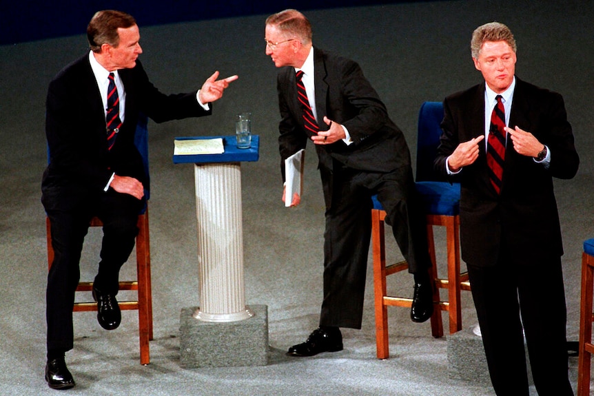 Three men in suits stand on a stage during a debate