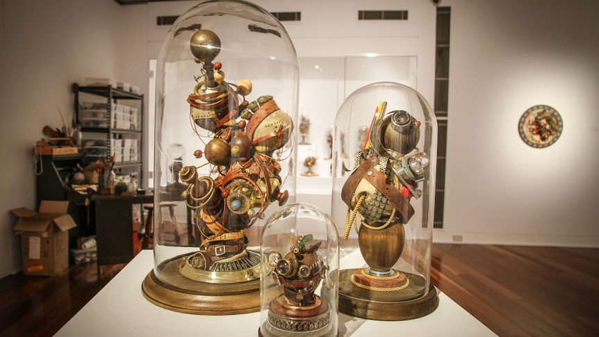 Three bell jars containing artworks sit on a table in an exhibition environment. 