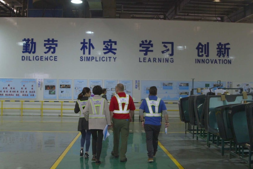 People in high vis vests in a factory with a big sign if Chinese and English that says 'diligence, simplicity, learning'