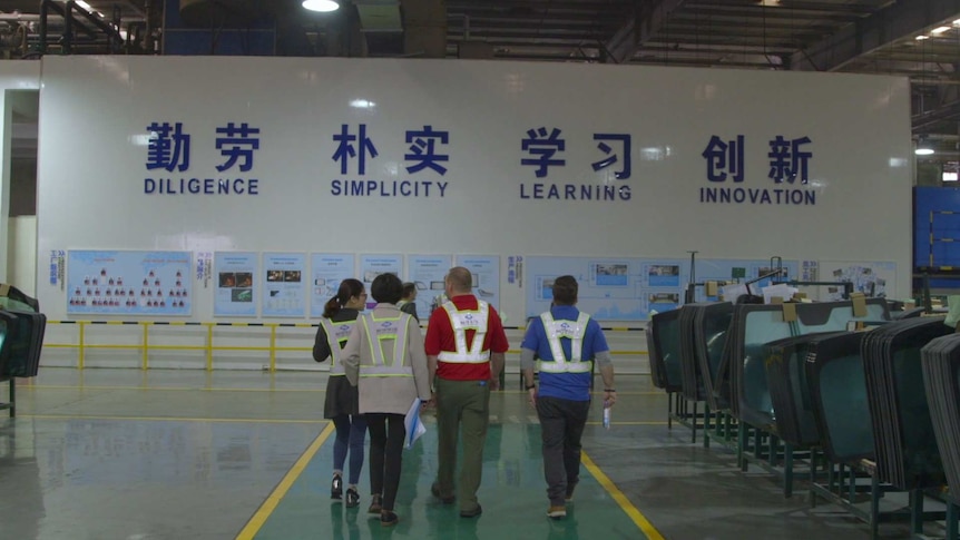People in high vis vests in a factory with a big sign if Chinese and English that says 'diligence, simplicity, learning'
