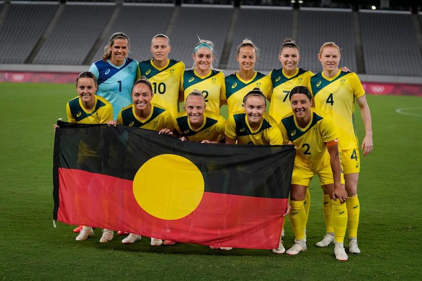 Australijska drużyna piłki nożnej kobiet, Matilda, pozuje z flagą Aborygenów przed swoim pierwszym meczem na Igrzyskach Olimpijskich w Tokio.