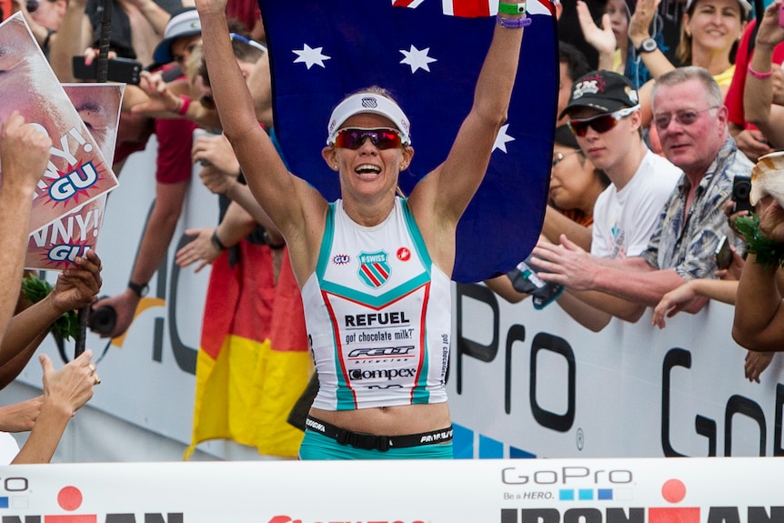 Mirinda Carfrae raises her hands and holds an Australian flag behind her
