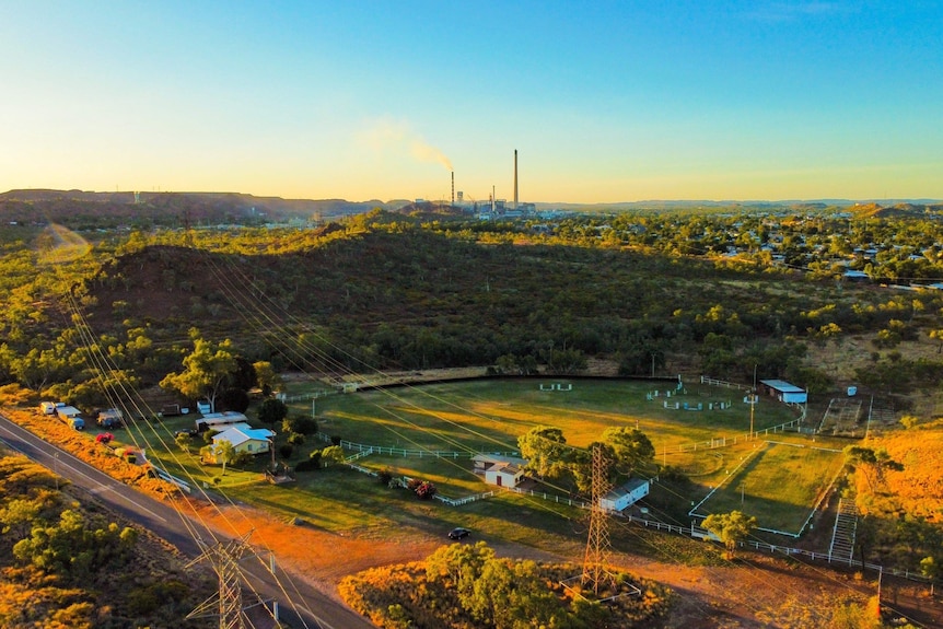An aerial view of an outback city shows a green sports oval and red dirt with the mining stack on the horizon