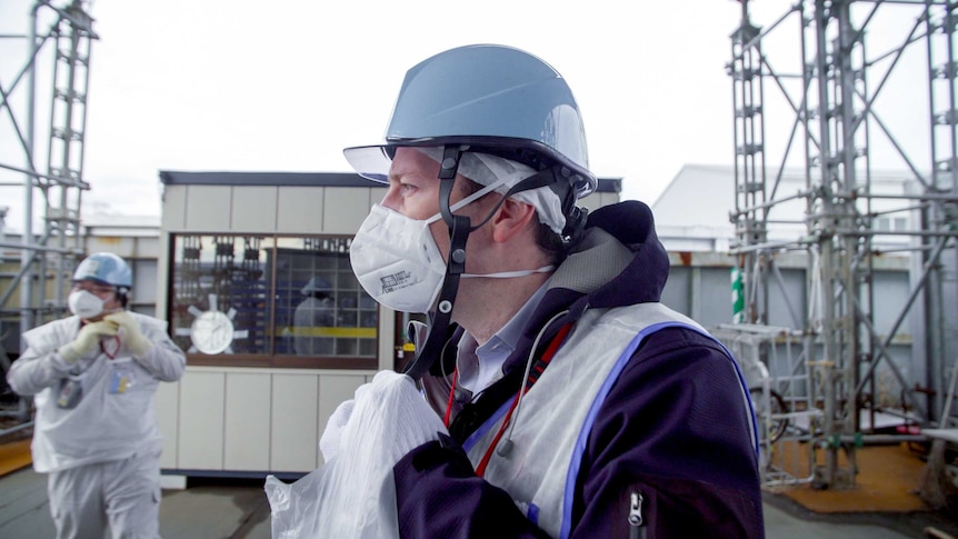 A man in a helmet, face mask and vest stands outside a nuclear plant