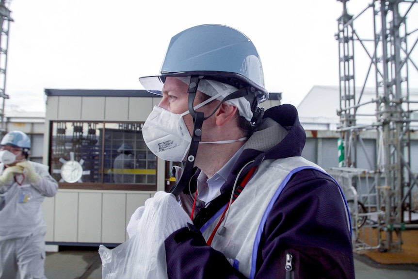 A man in a helmet, face mask and vest stands outside a nuclear plant