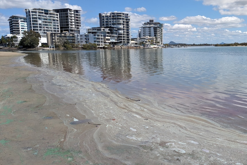 a large amount of brown liquid washing up on the beach with holiday apartments in the background.