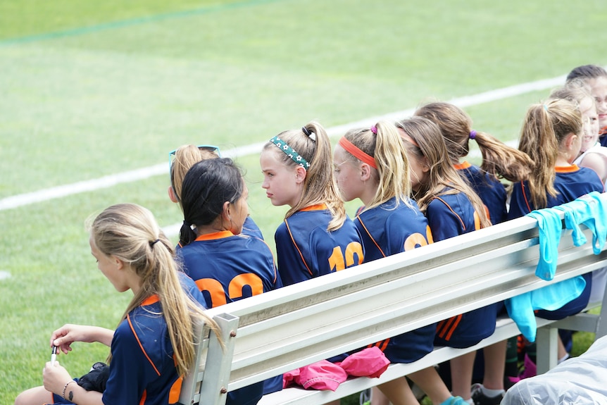 Girls in a sport uniform sit in a row on a silver bench.