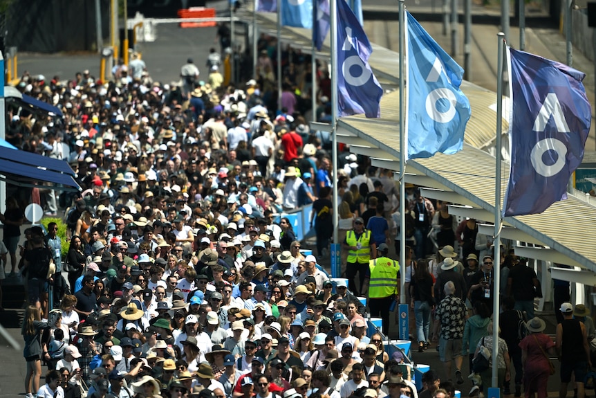 A large crowd of people moves past a train station. Australian Open flags are visible in the sky