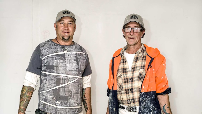 Two men in paint stained clothes and matching baseball caps hold buckets of paint.