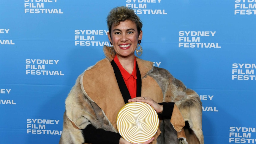 Director, writer and actor Kylie Bracknell stands in front of sydney film festival press wall wearing a kangaroo skin cloak