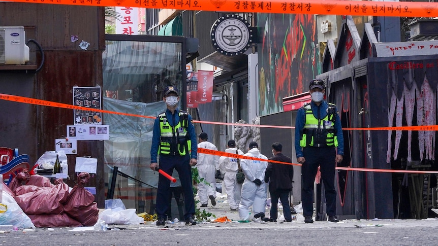 Two guards in mask stand before red tape as a group in white suits walk into a laneway.