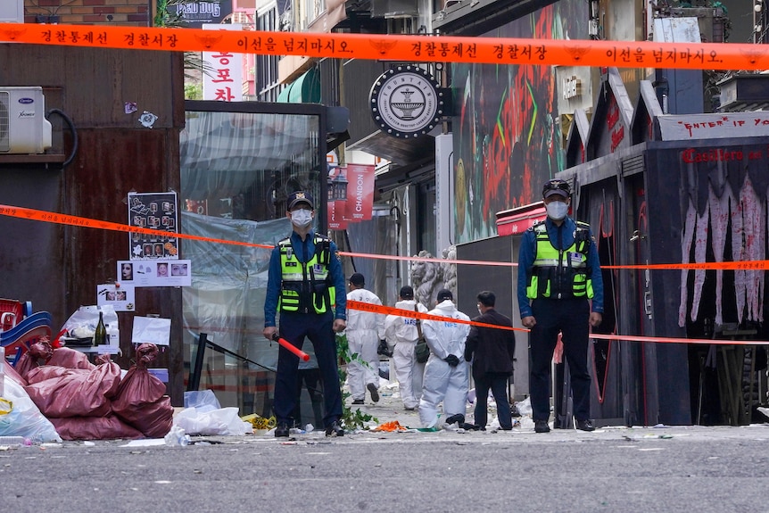 Two guards in mask stand before red tape as a group in white suits walk into a laneway.