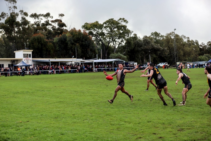 A player kicks a football under pressure as other players and spectators watch on at a country footy ground 
